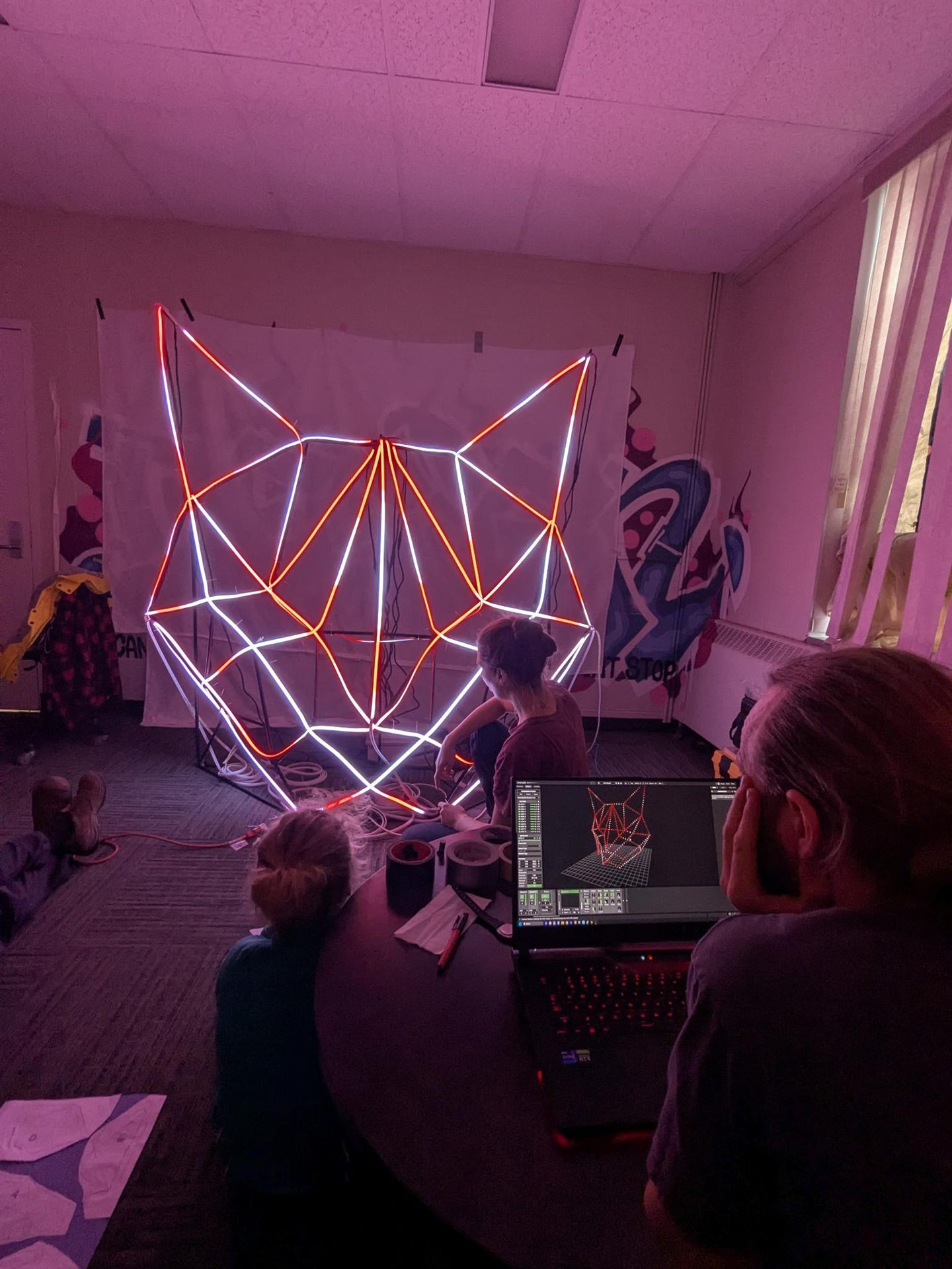 The assembled frame lit red and white in the workshop while one person wires it and another works on the 3D model on a laptop.