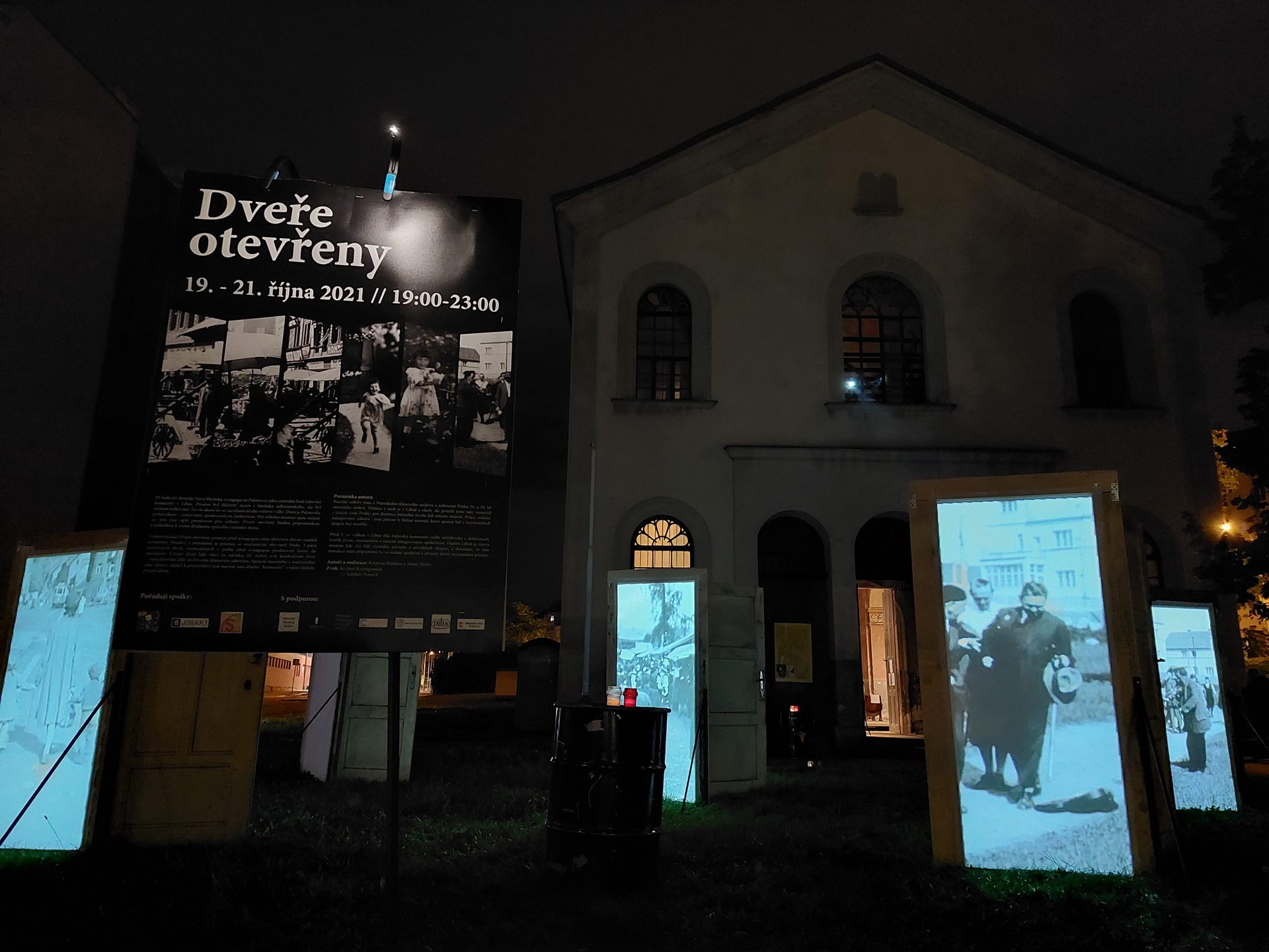 The New Liběn synagogue at night, a banner reading “Dveře otevřeny” beside the lit doors on the lawn.