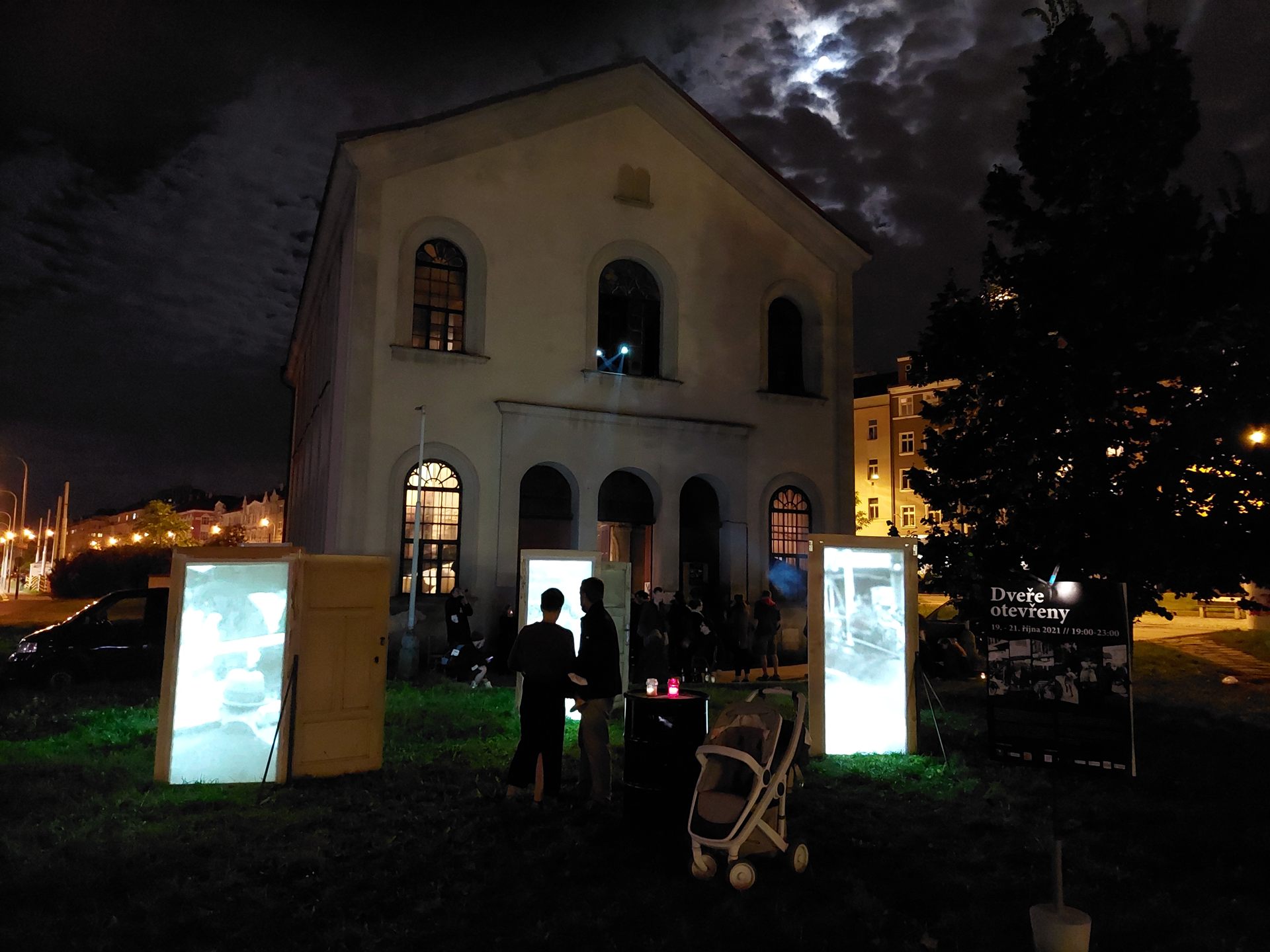 The synagogue under dramatic night clouds, the doors lit on the lawn in front of it.