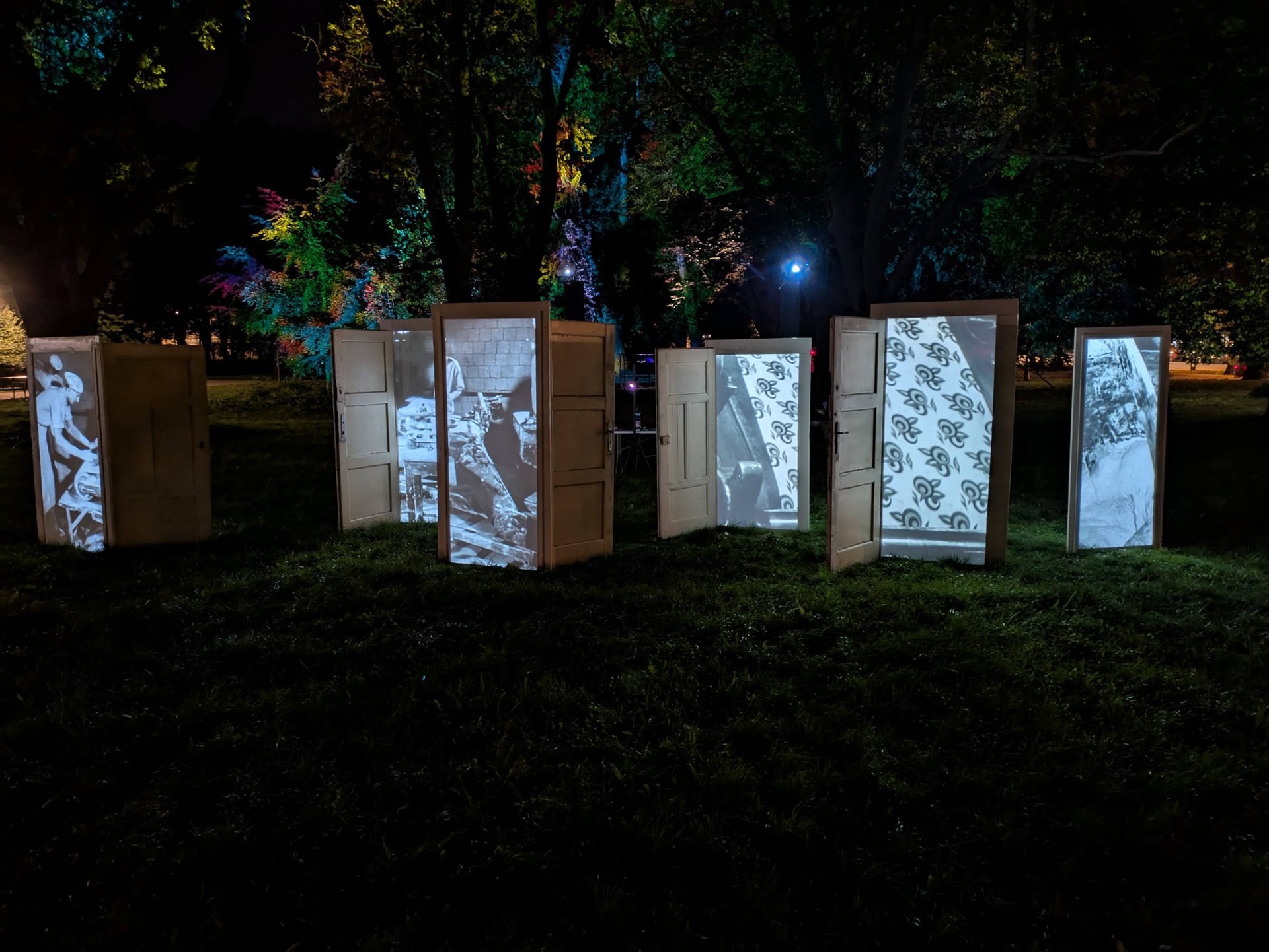 Six open doors standing in a row on grass at night, each projecting archival film footage.