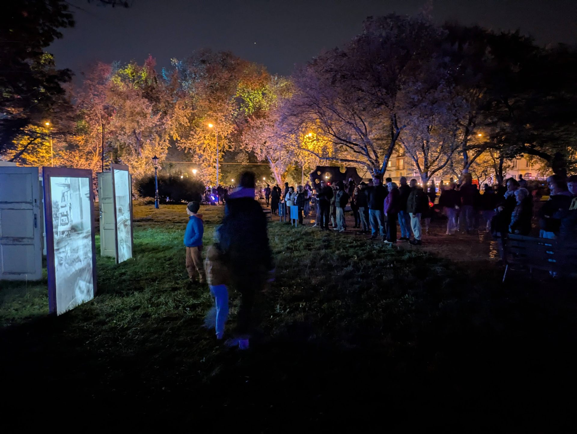 A crowd gathered under autumn trees lit in orange, one of the doors glowing at the left.