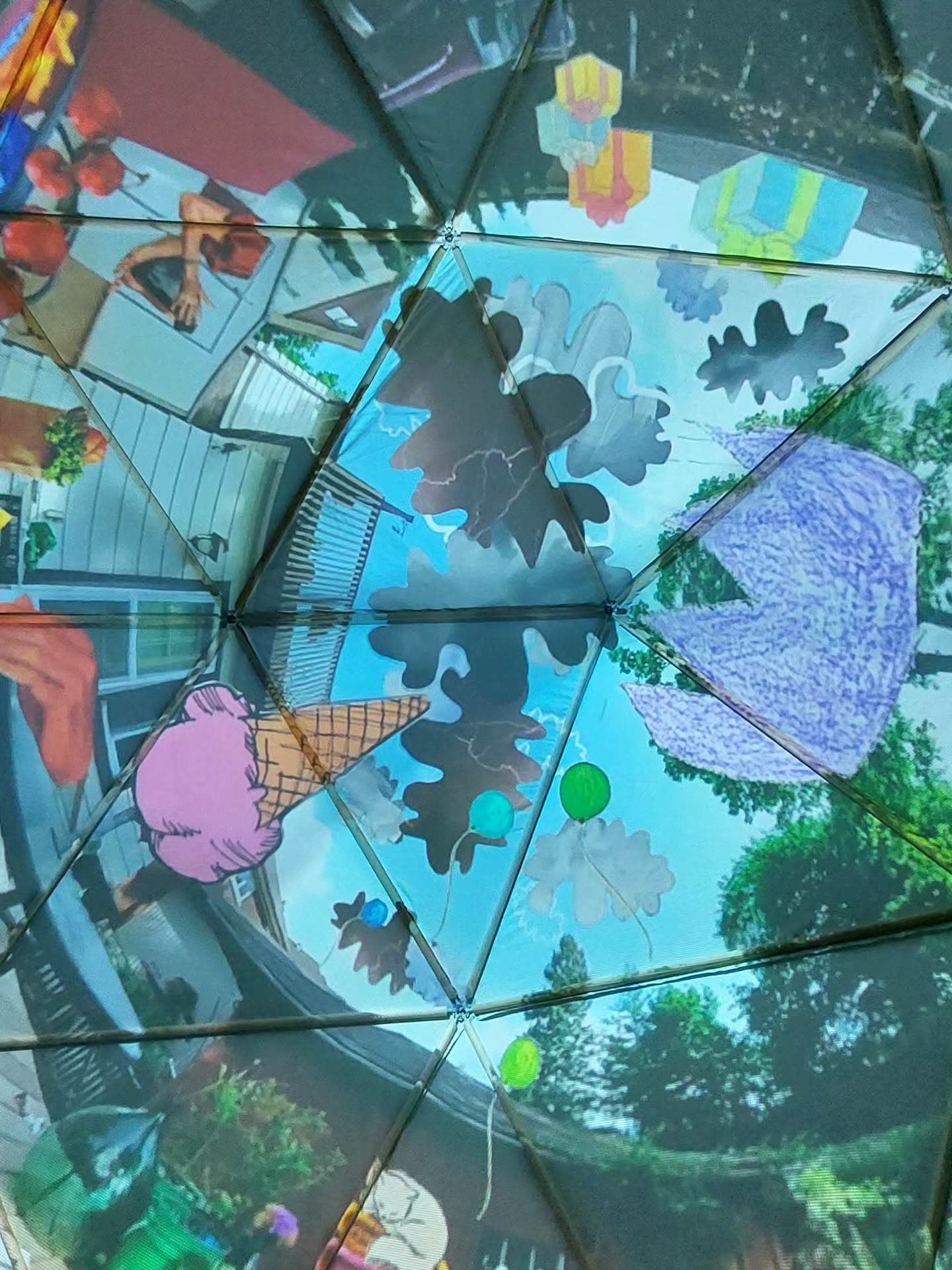 Looking up inside the geodesic dome, the triangular panels carrying a projected collage of a suburban street, storm clouds, balloons and an ice cream cone against blue sky.