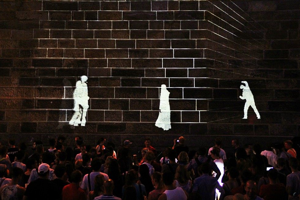 Three white projected figures walking across the stone wall above the audience.